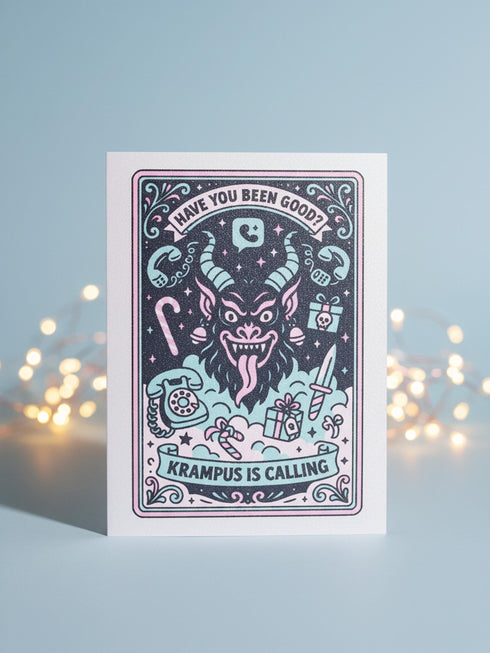A vertical 5x7 holiday card featuring a retro tattoo style illustration of Krampus in pastel pink, teal, and black ink on shimmery silver pearlescent paper. The central horned demon has a long tongue and is surrounded by vintage rotary phones, daggers, candy canes, and skull gifts. Top text asks Have You Been Good while the bottom banner reads Krampus Is Calling. The interior text reads Let's be honest... Being good is overrated. Merry Christmas.