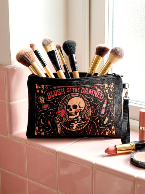 A rectangular gothic makeup bag made of black polyester with a visible woven linen-like crosshatch texture. The central illustration features a smiling human skull with flowing hair applying a pink heart-shaped blush to its cheek. Above the skull, the text BLUSH OF THE DAMNED appears. The design is framed by illustrations of lipstick tubes, mascara wands, bows. The bag includes a black zipper along the top edge and a removable black nylon wrist strap attached to the metal zipper pull.