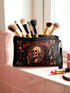 A rectangular gothic makeup bag made of black polyester with a visible woven linen-like crosshatch texture. The central illustration features a smiling human skull with flowing hair applying a pink heart-shaped blush to its cheek. Above the skull, the text BLUSH OF THE DAMNED appears. The design is framed by illustrations of lipstick tubes, mascara wands, bows. The bag includes a black zipper along the top edge and a removable black nylon wrist strap attached to the metal zipper pull.