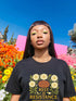 A black unisex t-shirt with a central front graphic. The design features the words "REST IS RESISTANCE" in a bold, gold, retro font. Above the main text, a serene, stylized face with closed eyes is framed by six cream-colored morning glory flowers and green leaves in a folk art, botanical style. Below, smaller text reads "SOFTNESS IS STRATEGY. SLEEP IS PROTEST." This alternative fashion, activist graphic tee is
