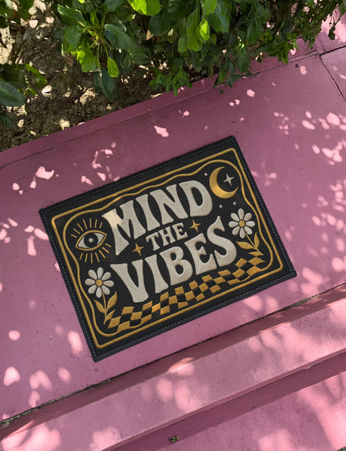 A rectangular floor mat with a dark charcoal black background. The center features the phrase 'MIND THE VIBES' in stylized, wavy cream and golden-mustard retro typography. The text is surrounded by symmetrical motifs: a golden evil eye with sun rays on the top left, a golden crescent moon with a four-point star on the top right, and two cream daisy flowers with golden stems at the bottom corners. The bottom edge has a wavy golden checkerboard pattern. A faux-stitch border frames the art.