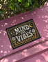A rectangular floor mat with a dark charcoal black background. The center features the phrase 'MIND THE VIBES' in stylized, wavy cream and golden-mustard retro typography. The text is surrounded by symmetrical motifs: a golden evil eye with sun rays on the top left, a golden crescent moon with a four-point star on the top right, and two cream daisy flowers with golden stems at the bottom corners. The bottom edge has a wavy golden checkerboard pattern. A faux-stitch border frames the art.
