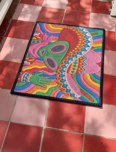 A rectangular doormat featuring a vibrant 2D flat print designed to look like textured yarn embroidery. The central graphic showcases a bright green alien with large black eyes, wearing a pink neck bandana and a highly decorated red, pink, and blue sombrero cowboy hat. The alien has one green hand raised in a friendly wave. The background is a psychedelic pattern of wavy, flowing rainbow stripes in yellow, orange, red, pink, blue, and green. The mat has a sleek black outer edge trim.