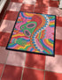 A rectangular doormat featuring a vibrant 2D flat print designed to look like textured yarn embroidery. The central graphic showcases a bright green alien with large black eyes, wearing a pink neck bandana and a highly decorated red, pink, and blue sombrero cowboy hat. The alien has one green hand raised in a friendly wave. The background is a psychedelic pattern of wavy, flowing rainbow stripes in yellow, orange, red, pink, blue, and green. The mat has a sleek black outer edge trim.