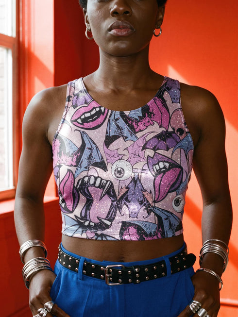 A sleeveless cropped tank top made from stretchy performance knit fabric. The all-over print features a dense, scattered pattern of pastel goth and creepy cute motifs over a light background. Graphics include pink and blue bats in flight, floating eyeballs with blue irises, pink skulls, and glossy vampire lips with sharp white fangs and tongues sticking out. The neckline is a high scoop and the armholes are curved. The fabric comes in either a matte finish or a sparkly silver glitter foil.