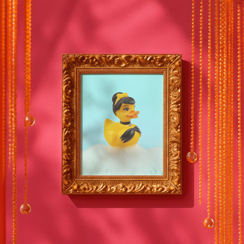 A chic rubber duck styled like Audrey Hepburn’s Holly Golightly, wearing a high bun, yellow bow, black gloves, and a sparkly choker. The duck floats in a pile of foam and is framed in white on a bubbly bathroom shelf with a red tile backdrop and soap dispenser.