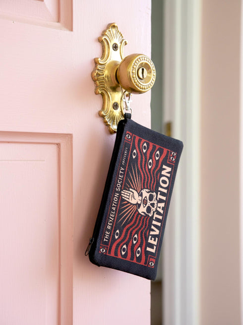 A rectangular makeup bag featuring a vibrant psychedelic print with gothic elements. The fabric texture is clearly visible showing a woven linen-like polyester grain. A black zipper runs across the top edge with a black detachable nylon wrist strap clip on the side, showcasing the approximately nine by six inch dimensions. The print includes high contrast neon colors and trippy swirling patterns typical of maximalist decor.