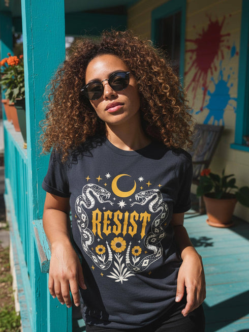 A unisex short sleeve graphic t shirt in dark charcoal black featuring a highly detailed symmetrical medallion print on the chest. In the center, the word RESIST is printed in a bold yellow gothic blackletter font. Two coiled white snakes with fine black scale details frame the text on the left and right. Above the text, a yellow crescent moon is flanked by white and yellow starbursts. Beneath, three yellow marigold flowers sit atop white stems and leafy foliage.