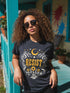 A unisex short sleeve graphic t shirt in dark charcoal black featuring a highly detailed symmetrical medallion print on the chest. In the center, the word RESIST is printed in a bold yellow gothic blackletter font. Two coiled white snakes with fine black scale details frame the text on the left and right. Above the text, a yellow crescent moon is flanked by white and yellow starbursts. Beneath, three yellow marigold flowers sit atop white stems and leafy foliage.