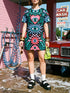 A short-sleeve, above-the-knee t-shirt dress with a true black background. The front features a vibrant, vertically stacked symmetrical graphic design. From top to bottom, the design includes a pink heart containing a green and blue eye, a large blue sunburst with a central eye, a red daisy flower with an eye flanked by pink and black checkered arches, and a pink mushroom cap with an eye. Swirling emerald green foliage, pink stars, and purple stars fill the negative space around the motifs.