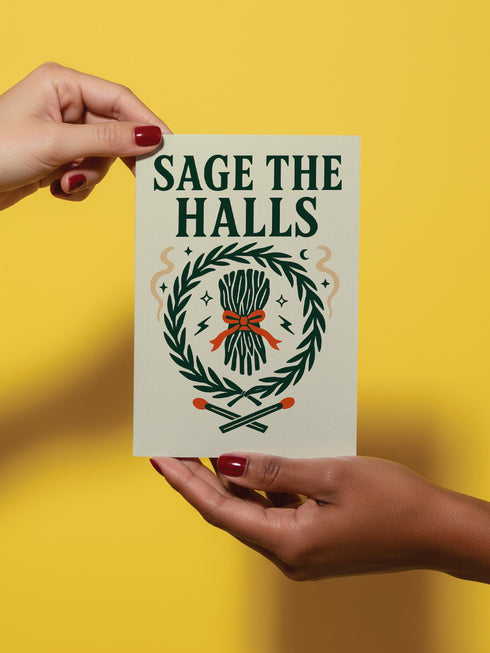 A detailed folk-art style illustration of a "Sage the Halls" holiday greeting card. The design features a bundle of sage smudge sticks tied with a red ribbon at the center of a dark green wreath. The wreath is accented with small crescent moons, stars, and smoke swirls. Below the wreath are two crossed matches with red tips. The card is printed on creamy beige paper with bold, dark green serif text. This funny spiritual and witchy Yule card is a unique pagan greeting for energy cleansing.