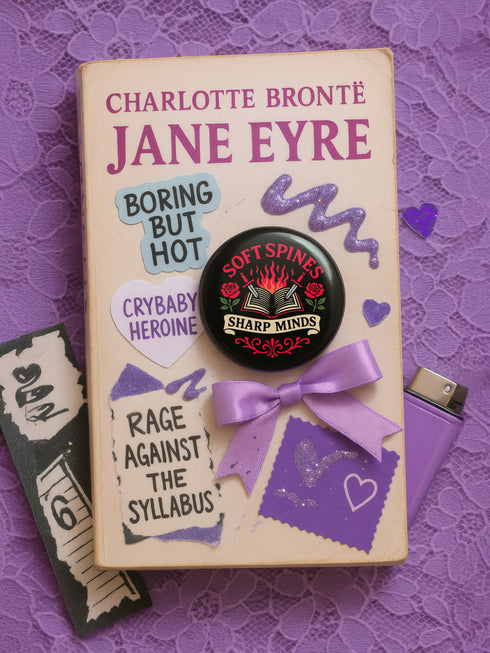 A round pinback button in black with a glossy mylar face and metal construction. The design, in a traditional tattoo style, features a central open book with flames rising from its pages. Two silver swords with black hilts are crossed over the book. On either side of the book is a red rose with a green stem and leaves. Below the book is a cream-colored banner with the words "SHARP MINDS" in a black serif font. Above the book, the words "SOFT SPINES" are written in a curved, red font.