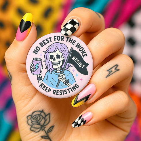 Honey Dazed Glossy round pink pinback button featuring a skeleton girl with purple hair holding a black flag that says “RESIST” and an energy drink can, with the phrases “No Rest for the Woke” and “Keep Resisting” in bold black letters.