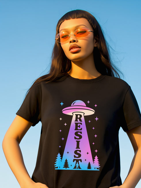 A person wearing a black unisex graphic t-shirt. The front of the shirt features a large, vibrant design with a retro-futuristic aesthetic. A classic flying saucer UFO, rendered in a pink and purple vaporwave gradient, hovers in the center. A beam of light descends from the UFO, containing the word 'RESIST' in a bold, uppercase font that transitions from purple to blue. Below the UFO, a stylized forest of neon blue and pink pine trees sits at the base of the design.