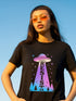 A person wearing a black unisex graphic t-shirt. The front of the shirt features a large, vibrant design with a retro-futuristic aesthetic. A classic flying saucer UFO, rendered in a pink and purple vaporwave gradient, hovers in the center. A beam of light descends from the UFO, containing the word 'RESIST' in a bold, uppercase font that transitions from purple to blue. Below the UFO, a stylized forest of neon blue and pink pine trees sits at the base of the design.