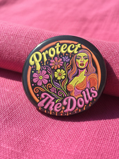 A circular black pinback button featuring the slogan Protect The Dolls in retro script. The design utilizes a high-fidelity trompe-l'œil print style that mimics the texture of raised thread embroidery with satin-stitch detailing. The artwork features vibrant hot pink, rust orange, and golden yellow motifs including drag queens with bouffant hair, 70s style daisies, lightning bolts, and a crescent moon with a face. The surface is smooth and glossy, creating a faux-textured illusion.