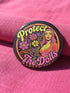 A circular black pinback button featuring the slogan Protect The Dolls in retro script. The design utilizes a high-fidelity trompe-l'œil print style that mimics the texture of raised thread embroidery with satin-stitch detailing. The artwork features vibrant hot pink, rust orange, and golden yellow motifs including drag queens with bouffant hair, 70s style daisies, lightning bolts, and a crescent moon with a face. The surface is smooth and glossy, creating a faux-textured illusion.