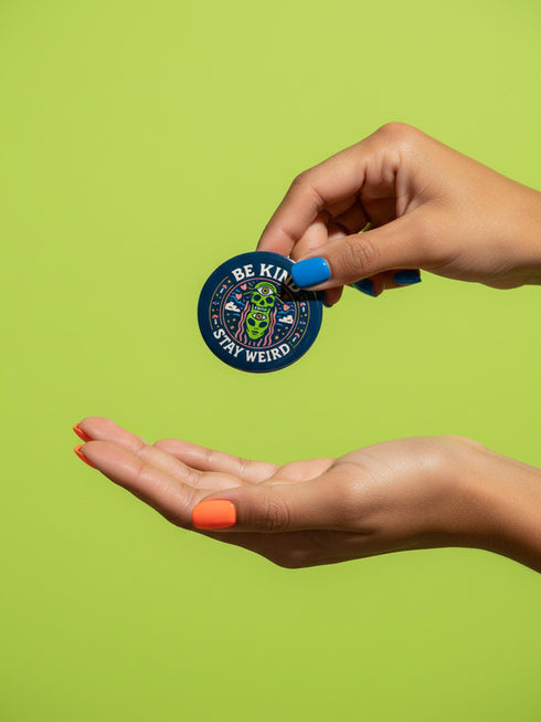 A round 1.75 inch pinback button featuring a retro psychedelic design on a dark navy blue background. The text reads BE KIND STAY WEIRD in vintage cream lettering around the border. The center illustration depicts a neon green skull stacked on top of a neon green alien head. Both the skull and alien have opening third eyes on their foreheads. Pink wavy lines frame the faces. The background is decorated with small colorful mushrooms, hearts, stars, and clouds. Glossy finish.
