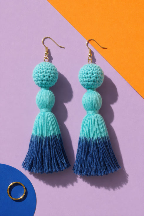 A pair of turquoise tassel earrings with gold hooks, displayed against a purple and orange background.