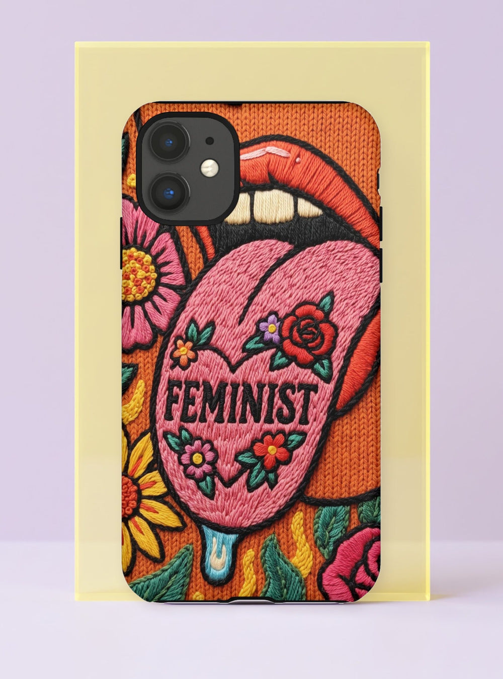 Dual layer matte phone case with a 2D print that mimics knit and embroidery texture. A retro open mouth with its tongue out sits over a pink heart stitched with the word FEMINIST in black letters, on an orange knit-look background filled with pink flowers, yellow sunflowers, red roses, purple and orange blooms, teal leaves, and a turquoise drip, all outlined in bold black. Smooth printed surface, open ports, camera cutout. Feminist maximalist phone accessory.