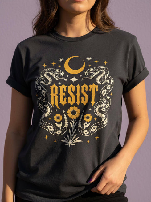 A unisex short sleeve graphic t shirt in dark charcoal black featuring a highly detailed symmetrical medallion print on the chest. In the center, the word RESIST is printed in a bold yellow gothic blackletter font. Two coiled white snakes with fine black scale details frame the text on the left and right. Above the text, a yellow crescent moon is flanked by white and yellow starbursts. Beneath, three yellow marigold flowers sit atop white stems and leafy foliage.