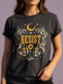 A unisex short sleeve graphic t shirt in dark charcoal black featuring a highly detailed symmetrical medallion print on the chest. In the center, the word RESIST is printed in a bold yellow gothic blackletter font. Two coiled white snakes with fine black scale details frame the text on the left and right. Above the text, a yellow crescent moon is flanked by white and yellow starbursts. Beneath, three yellow marigold flowers sit atop white stems and leafy foliage.