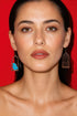 A woman with brown hair and a neutral expression, wearing large earrings with a bird design, against a red background.