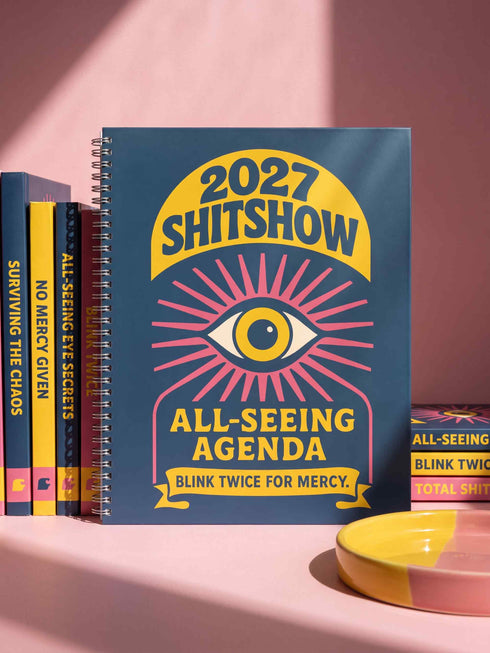A hardcover spiral planner for 2027 with a dark blue cover and retro 70s style graphics. The title reads "2027 SHITSHOW All-Seeing Agenda" in bold yellow and pink text. Below the title is a large, stylized all-seeing eye with pink rays emanating from it. At the bottom, a pink ribbon contains the text "BLINK TWICE FOR MERCY." The back cover is also dark blue with a yellow sun graphic and the text "Plans bloom. Deadlines hum. I write, I wander, then get it done. Keep receipts. Keep grace.