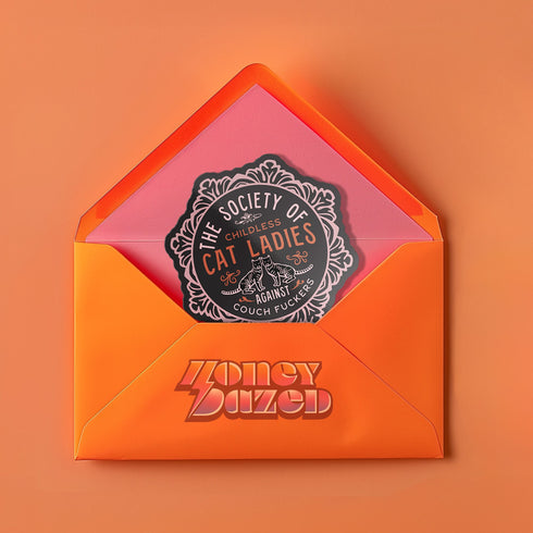 Round sticker with ornate border showing two cats and text "The Society of Childless Cat Ladies Against Couch Fuckers" in pink and purple versions on black background.