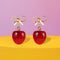 Gold bow earrings with dangling red cherry charms. Coquette-style jewelry perfect for soft girl and balletcore aesthetics. Handmade resin fruit earrings for summer style.
