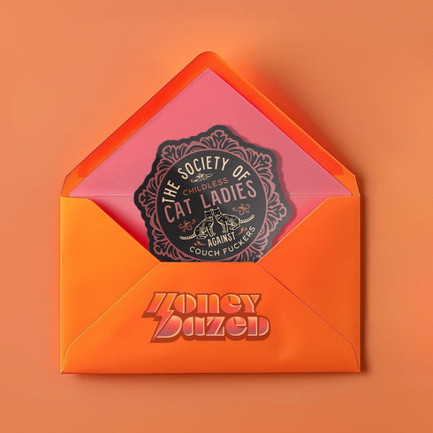 Round sticker with ornate border showing two cats and text "The Society of Childless Cat Ladies Against Couch Fuckers" in pink and purple versions on black background.