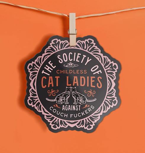 Round sticker with ornate border showing two cats and text "The Society of Childless Cat Ladies Against Couch Fuckers" in pink and purple versions on black background.