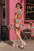 Short sleeve crew neck t-shirt dress in a relaxed, oversized straight cut, made of 100% polyester with a soft matte finish. An edge-to-edge all-over print on a cream background shows 60s daisies in red, yellow, and white set inside concentric rings of lime green, hot pink, orange, and royal blue, with magenta squares and red checkerboard accents. Retro 60s mod, flower power, rainbow maximalist alt fashion.