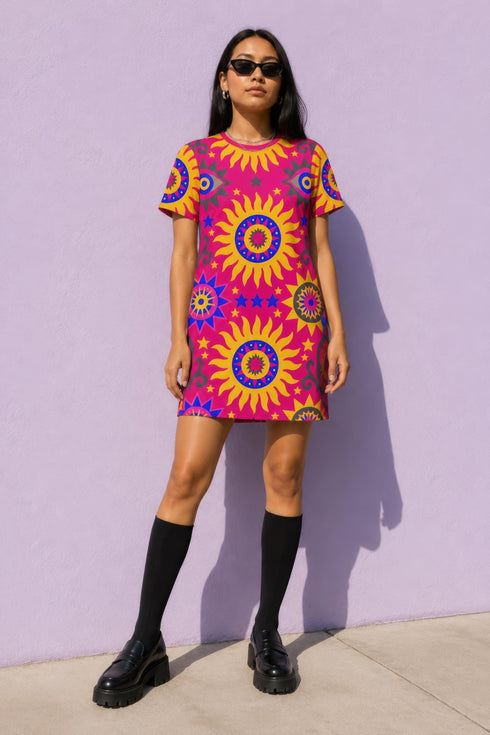 Short sleeve crew neck t-shirt dress in a relaxed, oversized straight cut, made of 100% polyester with a soft matte finish. An edge-to-edge all-over print shows yellow radial sunburst medallions with jagged rays on a hot pink background, layered blue and pink circular centers, rows of small blue stars, a partial eye near the top, and dark charcoal swirling vine filigree along the hem. Psychedelic 70s retro, celestial, whimsigoth maximalist alt fashion.