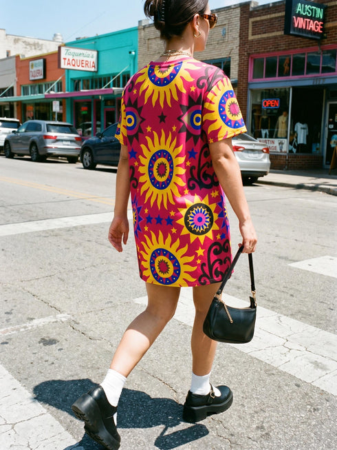 Short sleeve crew neck t-shirt dress in a relaxed, oversized straight cut, made of 100% polyester with a soft matte finish. An edge-to-edge all-over print shows yellow radial sunburst medallions with jagged rays on a hot pink background, layered blue and pink circular centers, rows of small blue stars, a partial eye near the top, and dark charcoal swirling vine filigree along the hem. Psychedelic 70s retro, celestial, whimsigoth maximalist alt fashion.