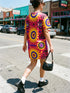Short sleeve crew neck t-shirt dress in a relaxed, oversized straight cut, made of 100% polyester with a soft matte finish. An edge-to-edge all-over print shows yellow radial sunburst medallions with jagged rays on a hot pink background, layered blue and pink circular centers, rows of small blue stars, a partial eye near the top, and dark charcoal swirling vine filigree along the hem. Psychedelic 70s retro, celestial, whimsigoth maximalist alt fashion.