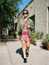 Short sleeve crew neck t-shirt dress in a relaxed, oversized straight cut, made of 100% polyester with a soft matte finish. An edge-to-edge all-over print shows yellow radial sunburst medallions with jagged rays on a hot pink background, layered blue and pink circular centers, rows of small blue stars, a partial eye near the top, and dark charcoal swirling vine filigree along the hem. Psychedelic 70s retro, celestial, whimsigoth maximalist alt fashion.