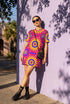 Short sleeve crew neck t-shirt dress in a relaxed, oversized straight cut, made of 100% polyester with a soft matte finish. An edge-to-edge all-over print shows yellow radial sunburst medallions with jagged rays on a hot pink background, layered blue and pink circular centers, rows of small blue stars, a partial eye near the top, and dark charcoal swirling vine filigree along the hem. Psychedelic 70s retro, celestial, whimsigoth maximalist alt fashion.