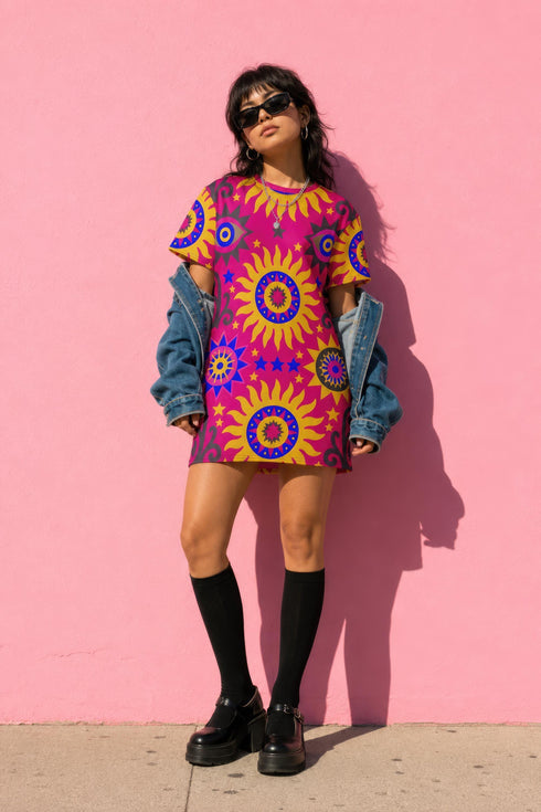 Short sleeve crew neck t-shirt dress in a relaxed, oversized straight cut, made of 100% polyester with a soft matte finish. An edge-to-edge all-over print shows yellow radial sunburst medallions with jagged rays on a hot pink background, layered blue and pink circular centers, rows of small blue stars, a partial eye near the top, and dark charcoal swirling vine filigree along the hem. Psychedelic 70s retro, celestial, whimsigoth maximalist alt fashion.