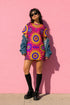 Short sleeve crew neck t-shirt dress in a relaxed, oversized straight cut, made of 100% polyester with a soft matte finish. An edge-to-edge all-over print shows yellow radial sunburst medallions with jagged rays on a hot pink background, layered blue and pink circular centers, rows of small blue stars, a partial eye near the top, and dark charcoal swirling vine filigree along the hem. Psychedelic 70s retro, celestial, whimsigoth maximalist alt fashion.