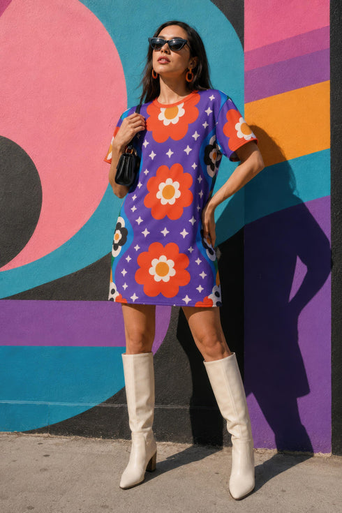 Short sleeve crew neck t-shirt dress in a relaxed, oversized straight cut, made of 100% polyester with a soft matte finish. An edge-to-edge all-over print on a medium purple background shows vertically stacked daisies with orange petals and yellow centers, scattered white four-point stars, and blue and green wavy side borders filled with small black and white daisies. Retro 70s floral, groovy maximalist alt fashion.