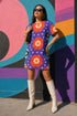 Short sleeve crew neck t-shirt dress in a relaxed, oversized straight cut, made of 100% polyester with a soft matte finish. An edge-to-edge all-over print on a medium purple background shows vertically stacked daisies with orange petals and yellow centers, scattered white four-point stars, and blue and green wavy side borders filled with small black and white daisies. Retro 70s floral, groovy maximalist alt fashion.