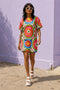 Short sleeve crew neck t-shirt dress in a relaxed, oversized straight cut, made of 100% polyester with a soft matte finish. An edge-to-edge all-over print on a cream background shows 60s daisies in red, yellow, and white set inside concentric rings of lime green, hot pink, orange, and royal blue, with magenta squares and red checkerboard accents. Retro 60s mod, flower power, rainbow maximalist alt fashion.