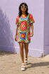 Short sleeve crew neck t-shirt dress in a relaxed, oversized straight cut, made of 100% polyester with a soft matte finish. An edge-to-edge all-over print on a cream background shows 60s daisies in red, yellow, and white set inside concentric rings of lime green, hot pink, orange, and royal blue, with magenta squares and red checkerboard accents. Retro 60s mod, flower power, rainbow maximalist alt fashion.