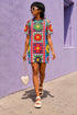 Short sleeve crew neck t-shirt dress in a relaxed, oversized straight cut, made of 100% polyester with a soft matte finish. An edge-to-edge all-over print shows 70s mod flowers in red, green, yellow, and purple set inside quilt-style patchwork panels of hot pink, royal blue, and lavender, divided by vertical stripes of red and pink polka dots. Retro 70s hippie, patchwork quilt, dopamine dressing maximalist alt fashion.