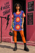 Short sleeve crew neck t-shirt dress in a relaxed, oversized straight cut, made of 100% polyester with a soft matte finish. An edge-to-edge all-over print on a medium purple background shows vertically stacked daisies with orange petals and yellow centers, scattered white four-point stars, and blue and green wavy side borders filled with small black and white daisies. Retro 70s floral, groovy maximalist alt fashion.