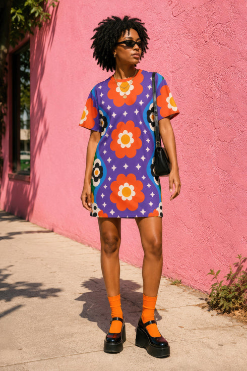 Short sleeve crew neck t-shirt dress in a relaxed, oversized straight cut, made of 100% polyester with a soft matte finish. An edge-to-edge all-over print on a medium purple background shows vertically stacked daisies with orange petals and yellow centers, scattered white four-point stars, and blue and green wavy side borders filled with small black and white daisies. Retro 70s floral, groovy maximalist alt fashion.