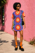 Short sleeve crew neck t-shirt dress in a relaxed, oversized straight cut, made of 100% polyester with a soft matte finish. An edge-to-edge all-over print on a medium purple background shows vertically stacked daisies with orange petals and yellow centers, scattered white four-point stars, and blue and green wavy side borders filled with small black and white daisies. Retro 70s floral, groovy maximalist alt fashion.