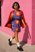 Short sleeve crew neck t-shirt dress in a relaxed, oversized straight cut, made of 100% polyester with a soft matte finish. An edge-to-edge all-over print on a medium purple background shows vertically stacked daisies with orange petals and yellow centers, scattered white four-point stars, and blue and green wavy side borders filled with small black and white daisies. Retro 70s floral, groovy maximalist alt fashion.