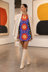 Short sleeve crew neck t-shirt dress in a relaxed, oversized straight cut, made of 100% polyester with a soft matte finish. An edge-to-edge all-over print on a medium purple background shows vertically stacked daisies with orange petals and yellow centers, scattered white four-point stars, and blue and green wavy side borders filled with small black and white daisies. Retro 70s floral, groovy maximalist alt fashion.